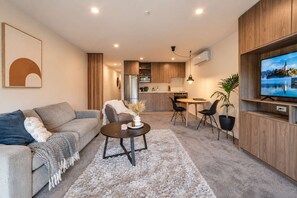 A modern living room with a sofa, coffee table, and chairs, a kitchen in the background, and a TV mounted on the wall.