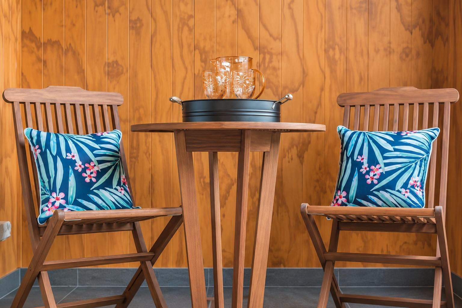 Two wooden chairs with floral cushions, a small round table, and a black pot with glassware on the table.