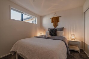 A bedroom with a large bed, a bedside table with a lamp, a window, and a wall-mounted macrame piece.