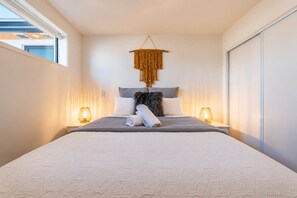 A modern bedroom with a large bed, two bedside lamps, a hanging macrame piece, and a view of the outside.