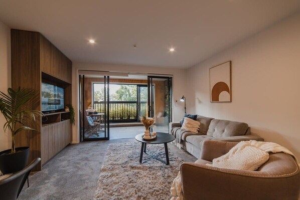 A modern living room with a sofa, a coffee table, and a TV. There is a sliding glass door leading to an outdoor area.