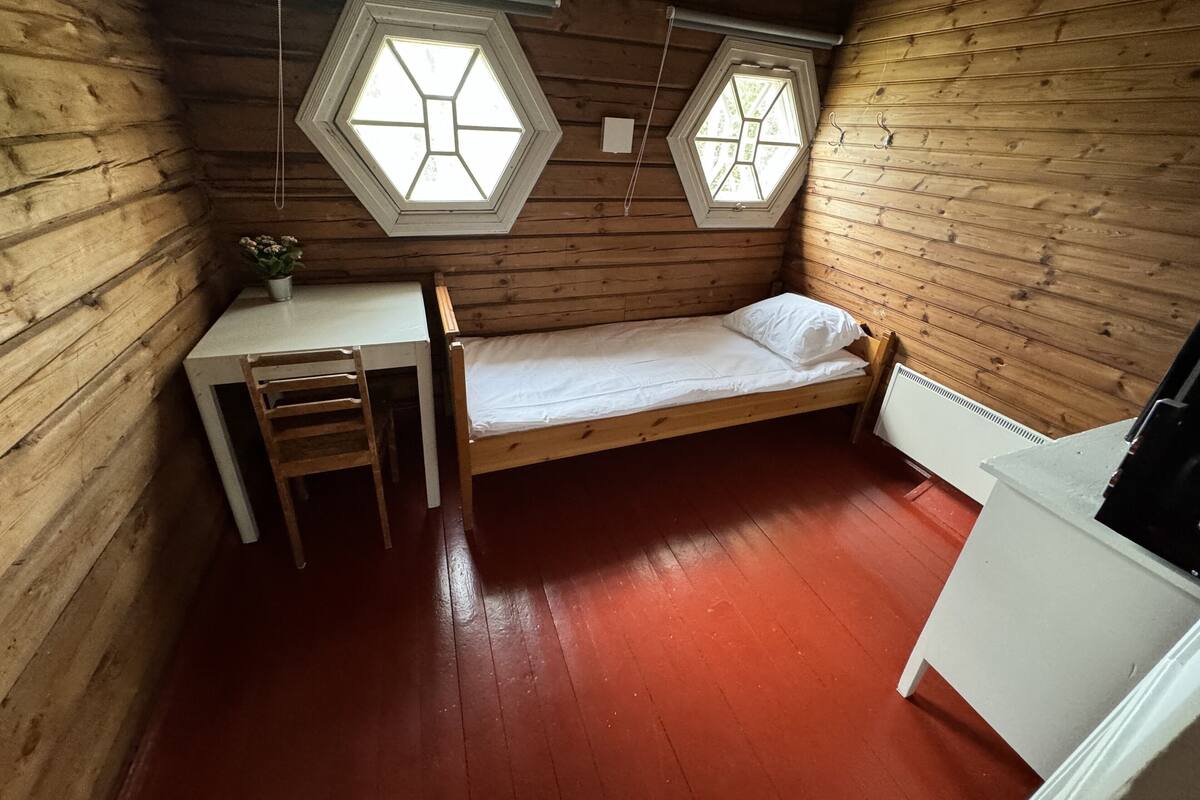 Une petite pièce en forme de cabane, avec des murs en bois, un lit simple, un bureau et deux fenêtres hexagonales.
