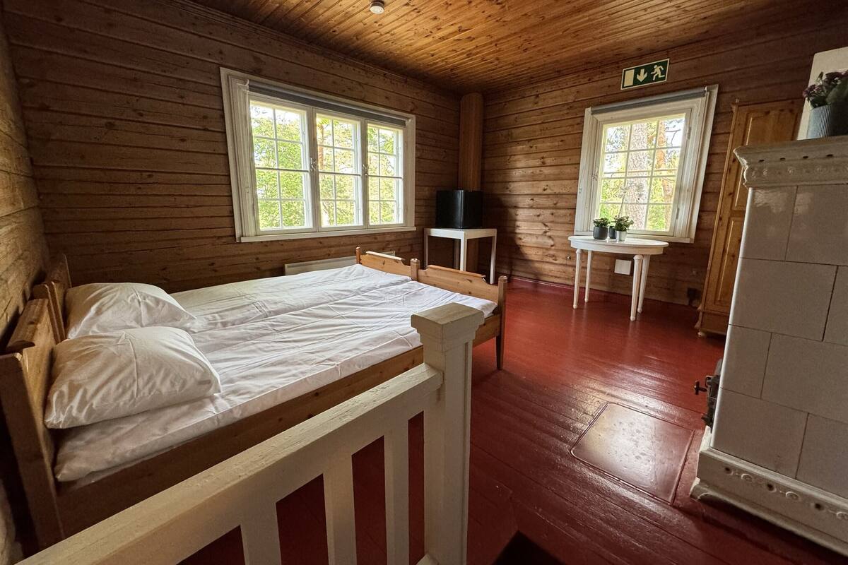 Une pièce de cabane en bois comprenant un lit, deux fenêtres, une petite table et un réfrigérateur.