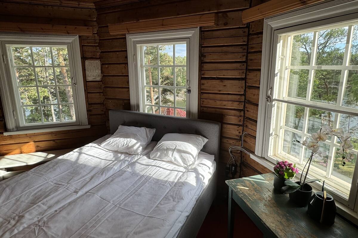 Une chambre dans une cabane en bois, avec un lit, deux fenêtres et une table ornée de plantes en pot.