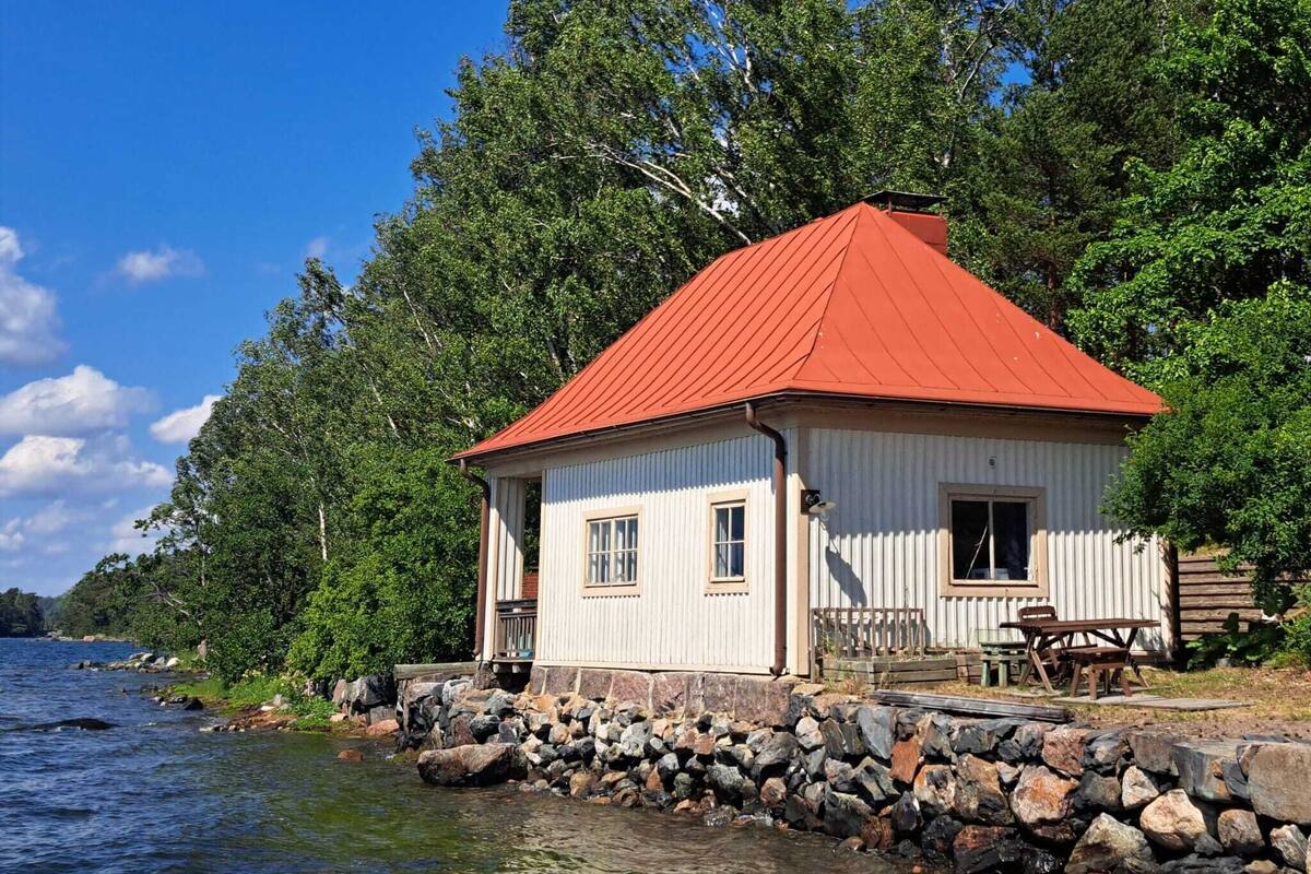 Une cabane blanche avec un toit rouge, située au bord d’un lac et entourée d’arbres.