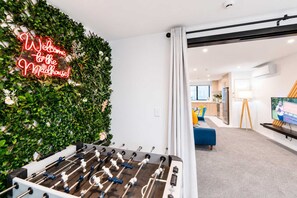 Ett rum med ett bordtennisspel, en neonreklamskylt som hänger på väggen, en soffa, en tv och ett köksområde i bakgrunden.