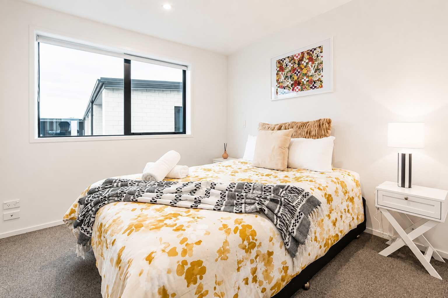 Habitación de hotel con una cama grande, una mesita de noche con lámpara, una ventana con vista a un edificio y una obra de arte en la pared.
