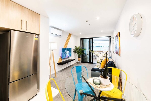 A compact living space with a kitchenette, a flat-screen TV, a dining area with a table and chairs, and a view through a sliding glass door.