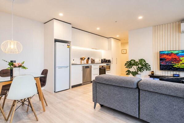 Un salon moderne avec un canapé gris, une table à manger avec des chaises, un coin cuisine doté de meubles blancs et un téléviseur à écran plat fixé au mur.