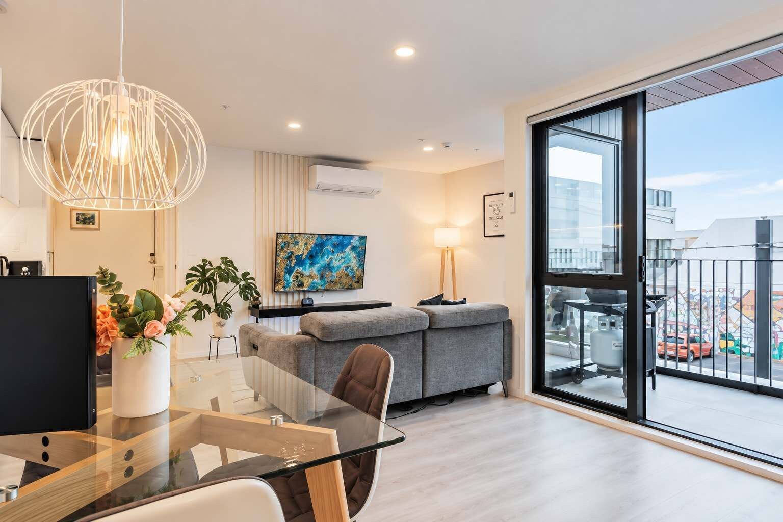 A modern living room with a glass dining table, grey sofa, and a large window with a city view.
