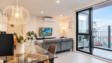 A modern living room with a glass dining table, grey sofa, and a large window with a city view.