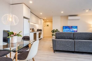 A modern living room with a grey sofa, a glass dining table with chairs, a kitchen area with white cabinets, and a flat-screen TV mounted on the wall.