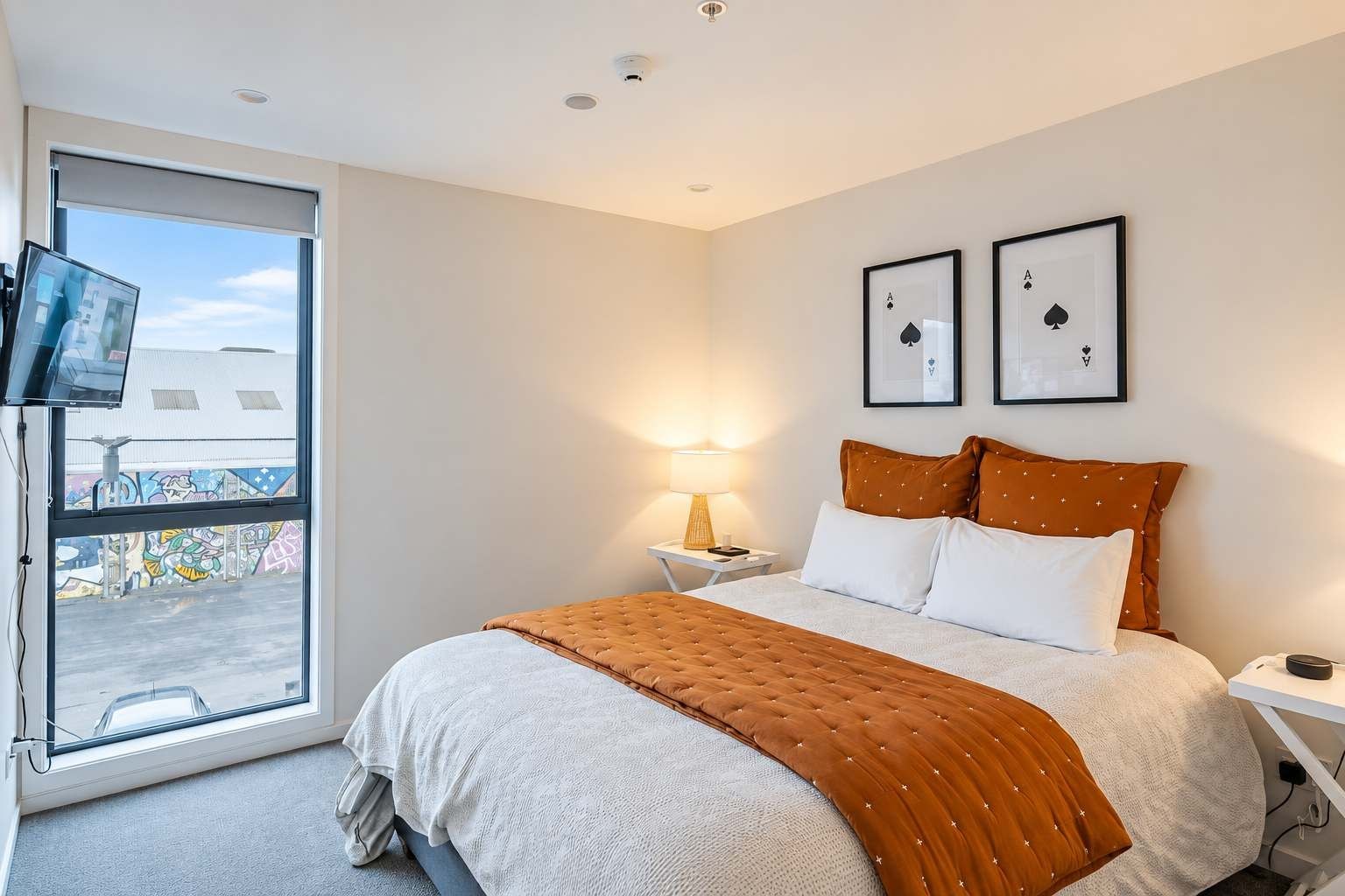 A hotel room with a bed, a TV, a window with a view of graffiti, and two framed playing cards on the wall.