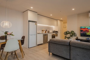 Una cocina moderna con armarios blancos, un comedor con mesa y sillas de vidrio, y un sofá gris en la sala.