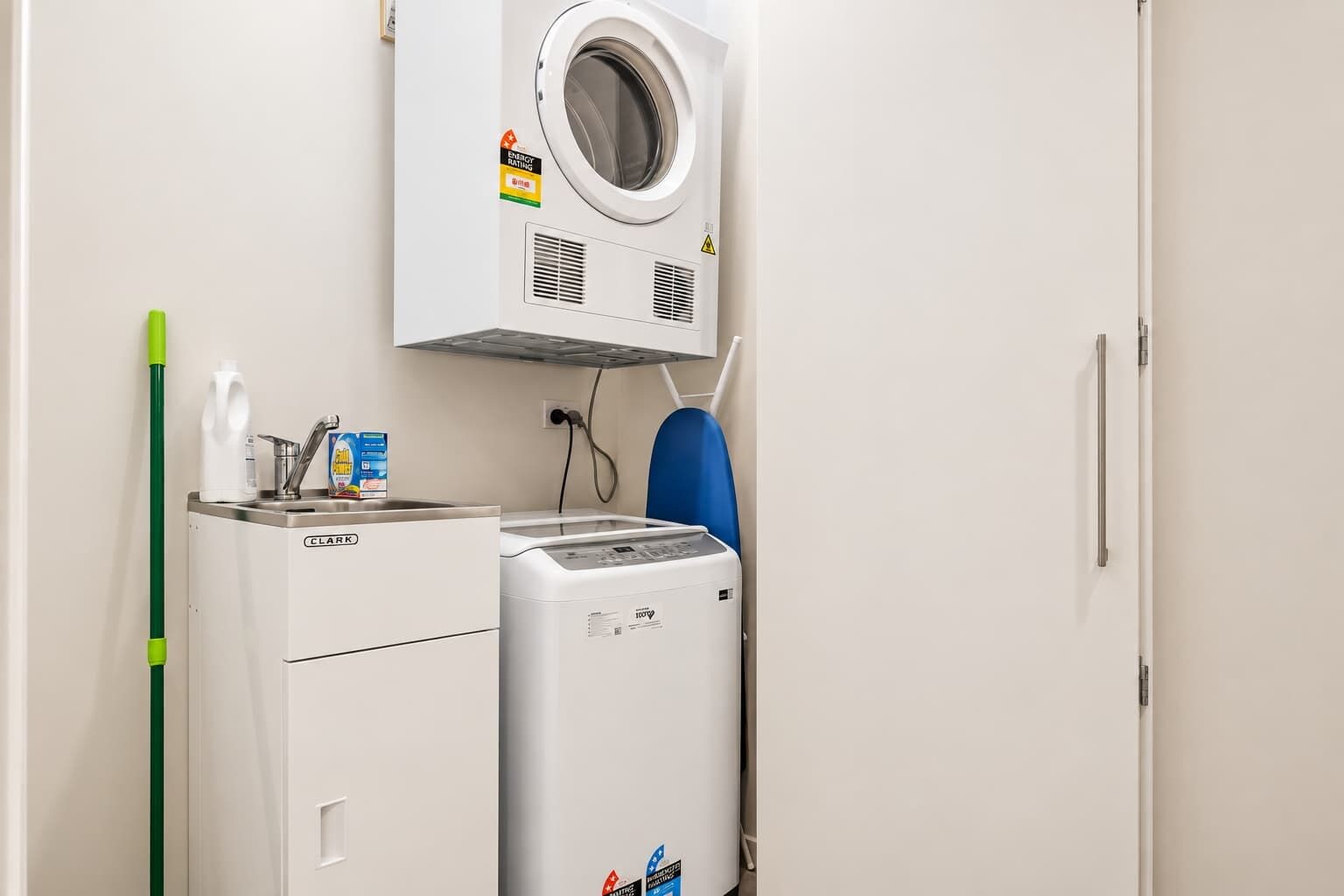 A laundry area with a front-loading washer and dryer, a green mop, and a bottle of detergent.