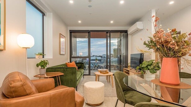 Ein modernes Wohnzimmer mit einem grünen Sofa, einem Ledersessel und einem Glastisch. Ein großes Fenster bietet Blick auf die Stadt, und auf einem Beistelltisch steht eine Pflanze.
