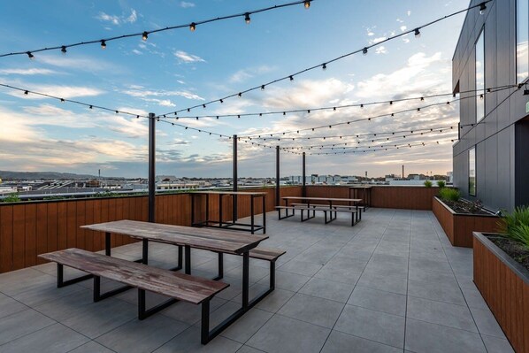 An outdoor patio with picnic tables and string lights against a cityscape backdrop.