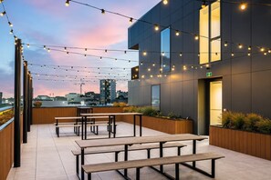 An outdoor terrace with wooden benches and string lights against a cityscape backdrop.