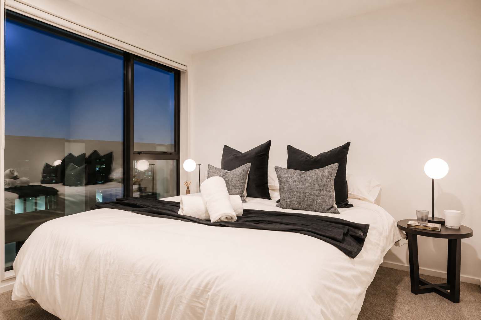 Ein Hotelzimmer mit einem großen Bett, einem Nachttisch mit Lampe und Blick auf die Stadt durch ein großes Fenster.