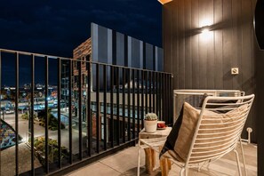 A balcony with a view of a cityscape at night, a white chair, a small table with a plant and a cup, and a wall-mounted light.