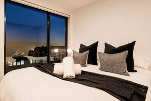A hotel room with a large bed, multiple pillows, a black throw blanket, and a view of the city at night through a large window.