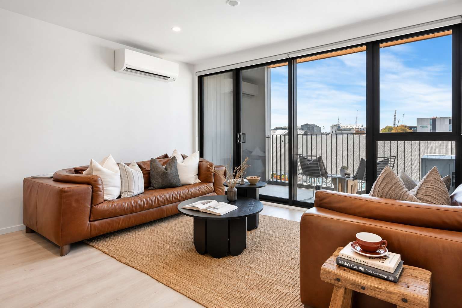 A modern living room with a brown leather sofa, a black coffee table, and a view of the city through large windows.