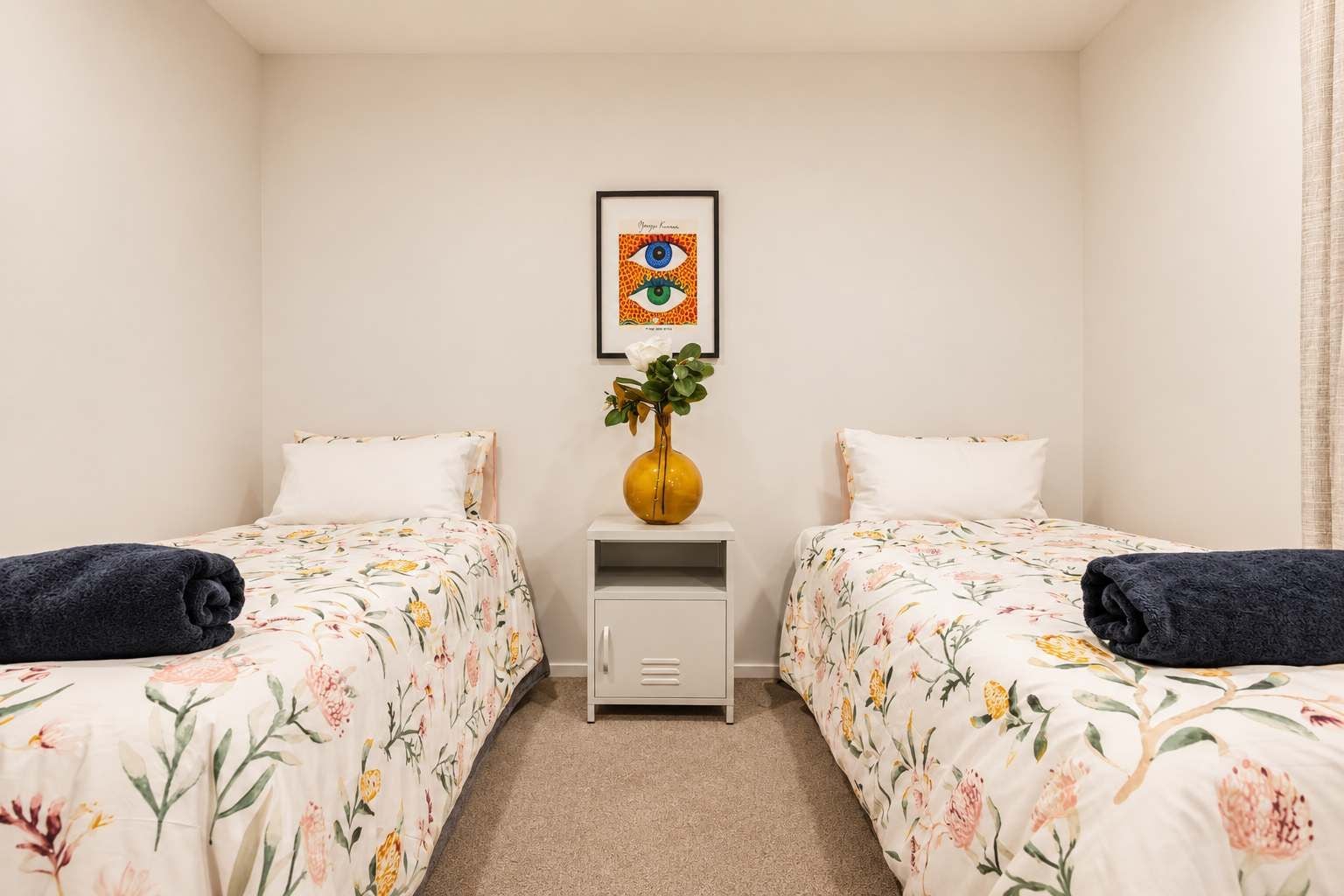Habitación de hotel con dos camas, una mesita de noche con un florero y un cuadro en la pared.