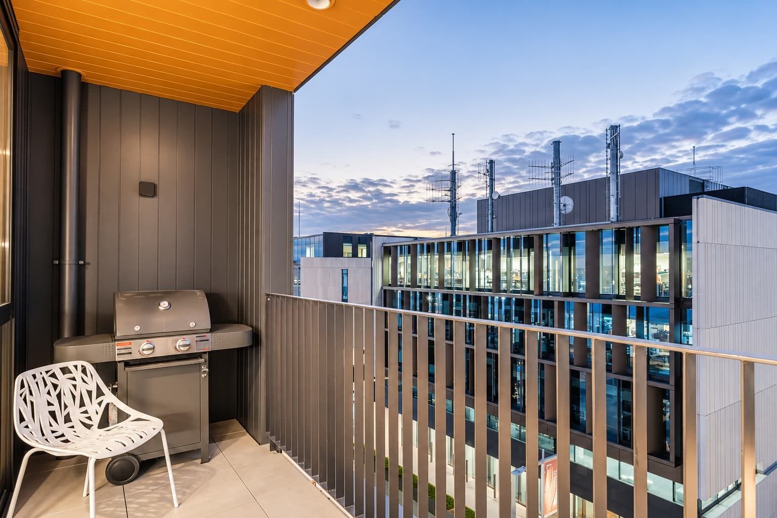Un balcón con una parrilla y una silla, con vistas a un edificio moderno de varios pisos y grandes ventanales.