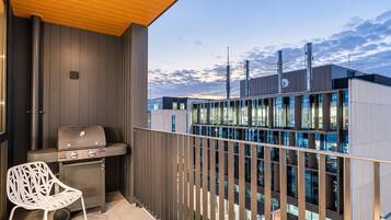 Un balcón con una parrilla y una silla, con vistas a un edificio moderno de varios pisos y grandes ventanales.
