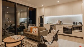 Uma sala de estar moderna com sofá, mesa de centro e poltrona aconchegante. Há uma cozinha equipada com geladeira, micro-ondas e fogão.
