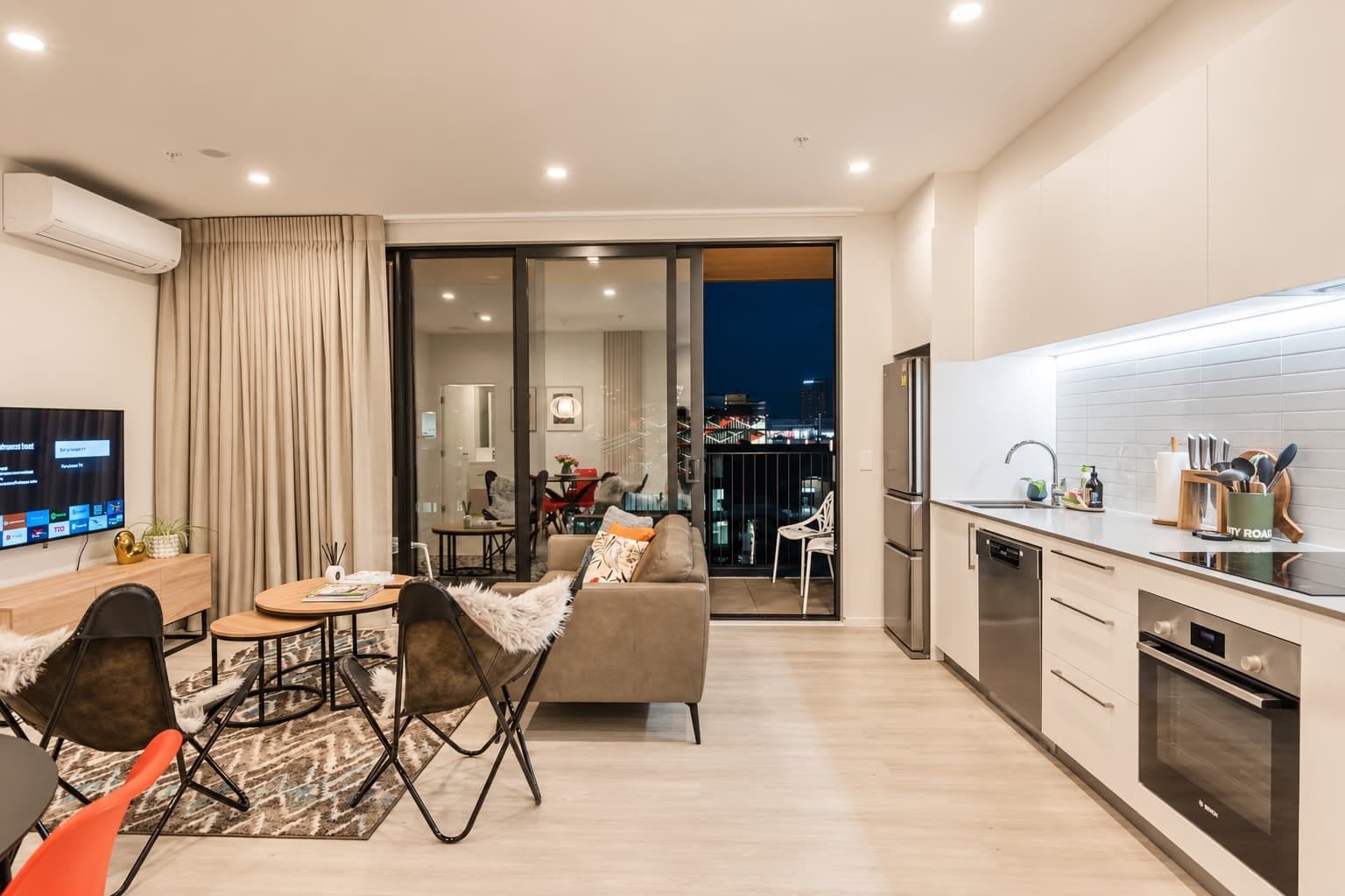 A modern apartment with a dining area, sofa, kitchen, and a balcony with a city view.