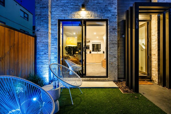 Una casa moderna con puerta de entrada de vidrio, un pequeño patio con sillas y un césped bien cuidado.