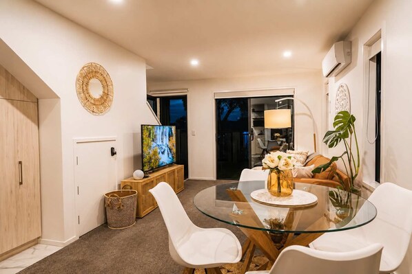A modern living room with a glass dining table, white chairs, a TV, and a sofa.