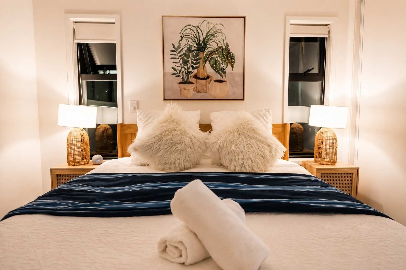 A neatly made bed with a blue blanket, two fluffy pillows, a rolled towel, and two bedside lamps on wooden nightstands.
