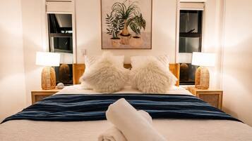 A neatly made bed with a blue blanket, two fluffy pillows, a rolled towel, and two bedside lamps on wooden nightstands.