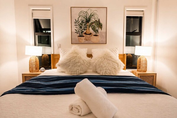 Una cama bien hecha con una manta azul, dos almohadas mullidas, una toalla enrollada y dos lámparas de noche sobre mesitas de madera.