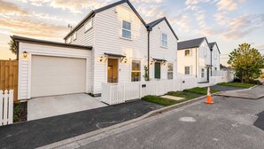 A residential street with white houses, a white picket fence, and a traffic cone on the road.