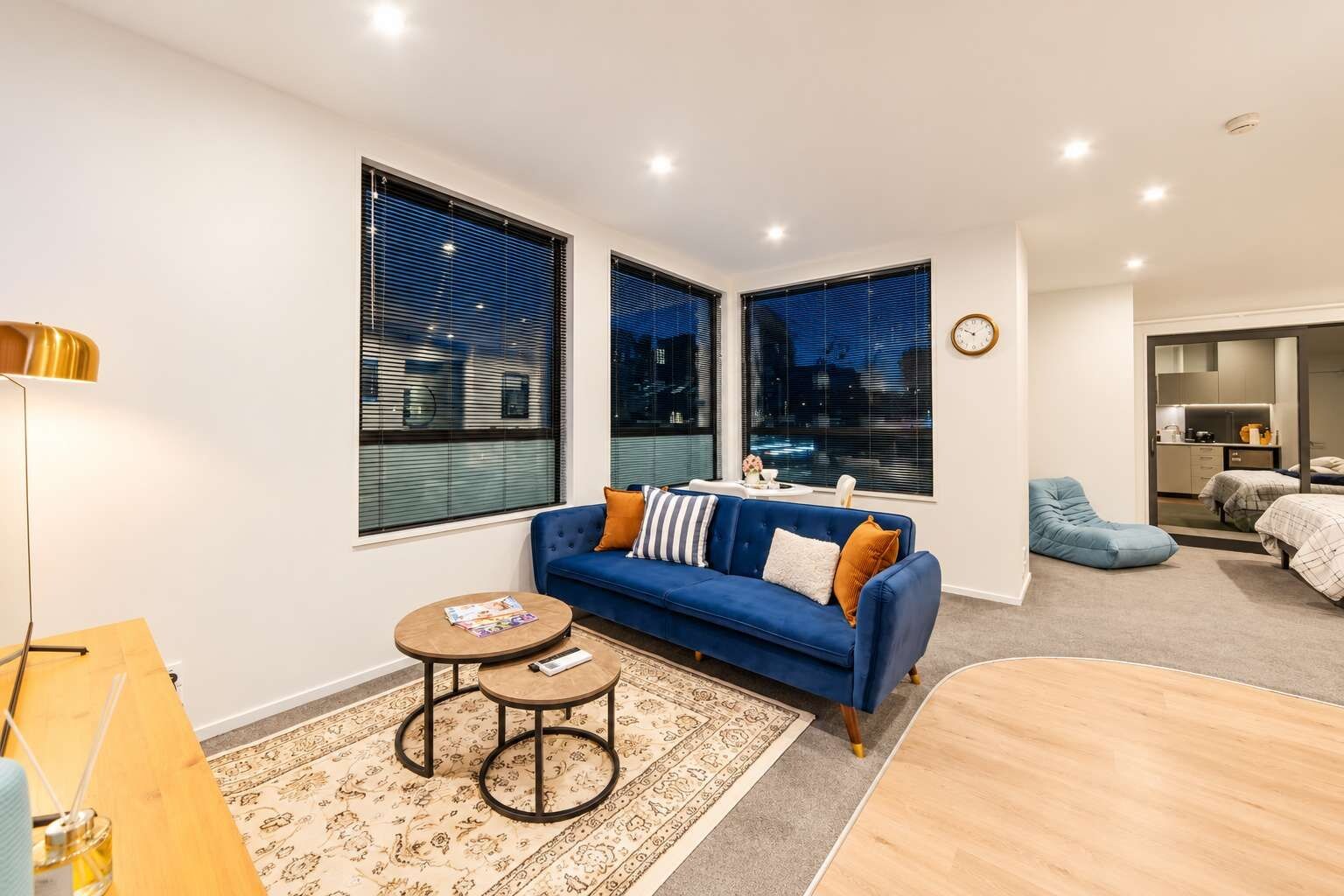 A modern living room with a blue sofa, a coffee table, and a rug. There are two windows with blinds, a clock on the wall, and a kitchenette in the background.