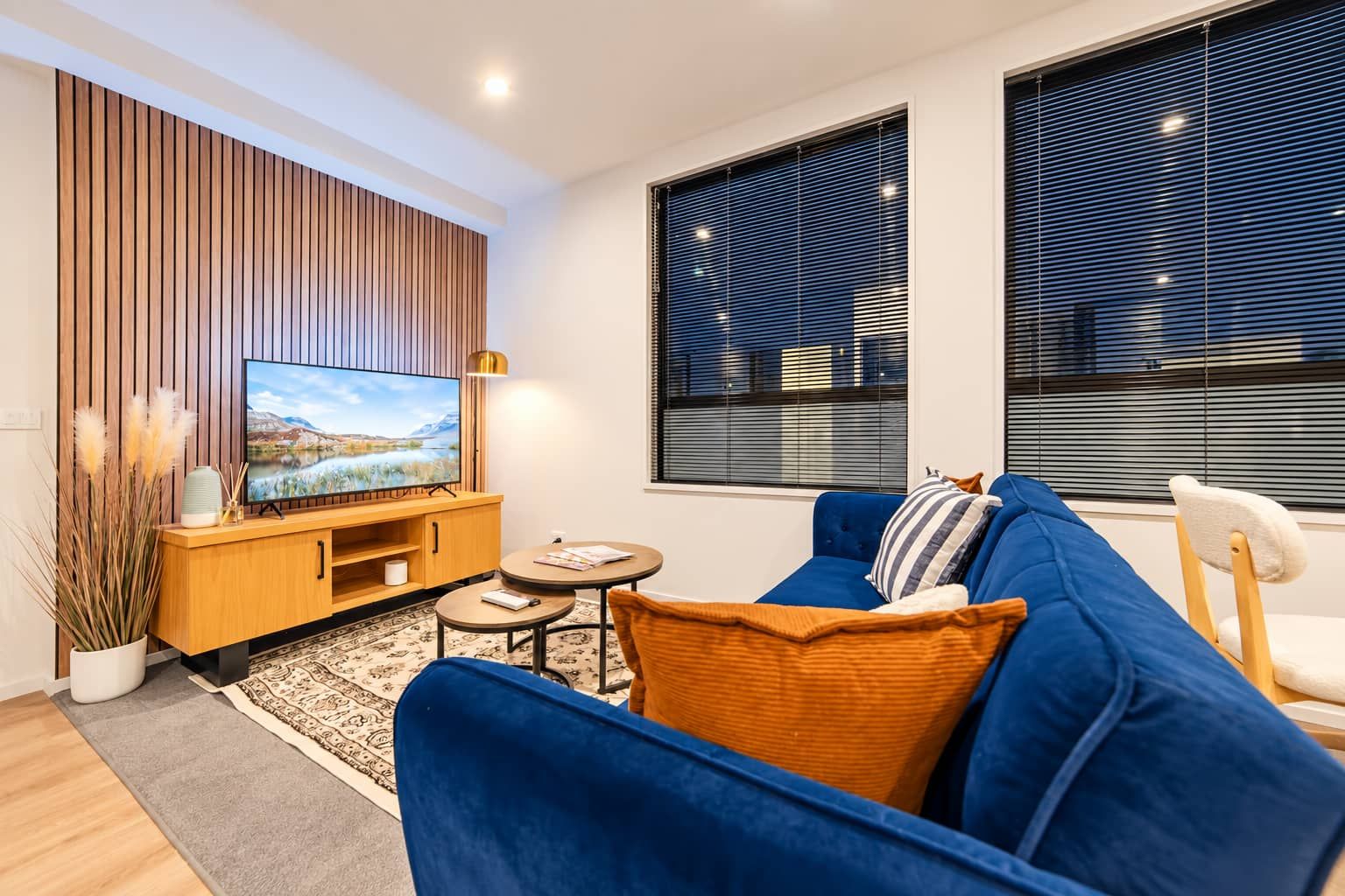 A modern living room with a flat-screen TV, a wooden entertainment unit, a blue sofa, a small round table, and a large window with blinds.