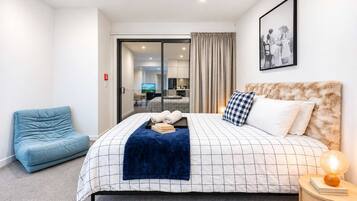 Une chambre d’hôtel avec un lit, un fauteuil bleu, une table de chevet avec une lampe, et une vue sur l’intérieur de l’hôtel.