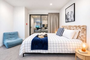 A hotel room with a bed, a blue armchair, a bedside table with a lamp, and a view of the hotel's interior. - Fitzgerald City Base | 1BR + Patio + WiFi (Christchurch)