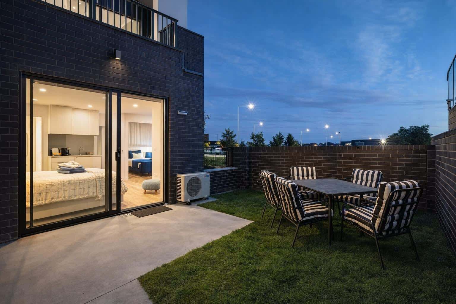 A modern building with a glass door entrance leading to a bedroom, a patio with a table and chairs, and a brick wall.