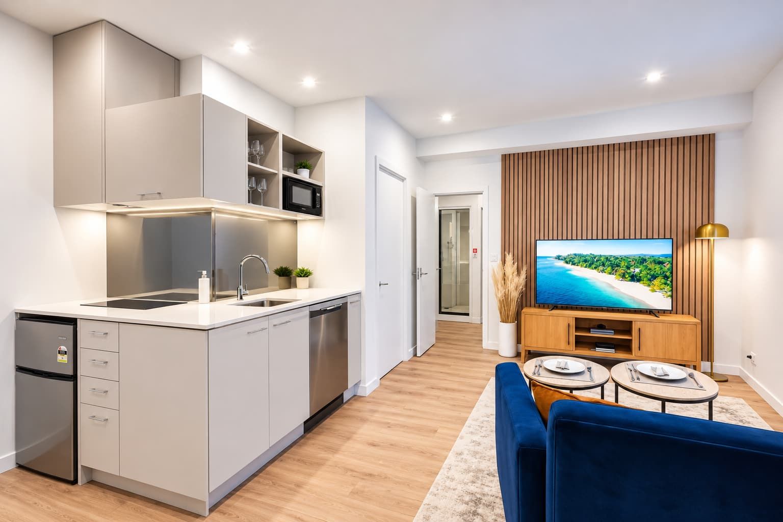 A modern kitchen with a built-in microwave, a sink, and a dishwasher. There is a dining area with a blue sofa and a TV displaying a beach scene.