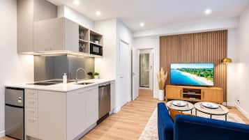 A modern kitchen with a built-in microwave, a sink, and a dishwasher. There is a dining area with a blue sofa and a TV displaying a beach scene.