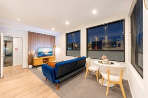 A modern living room with a blue sofa, a dining table with chairs, a TV on a wooden stand, and large windows with blinds. - Fitzgerald City Base | 1BR + Patio + WiFi (Christchurch)