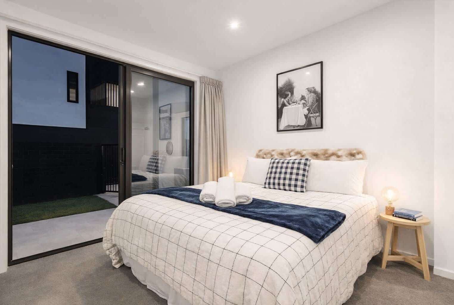 A bedroom with a bed, a bedside table with a lamp, a framed picture on the wall, and a sliding glass door leading to an outdoor area.