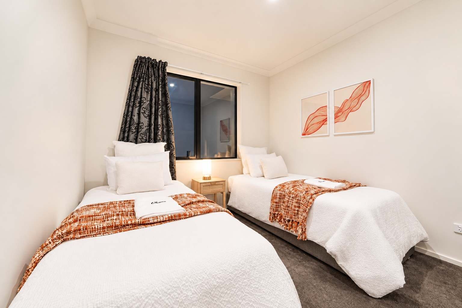 Una habitación de hotel con dos camas, una ventana con cortinas, una mesita de noche con lámpara y una obra de arte en la pared.