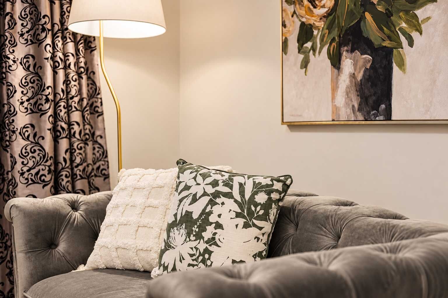 A tufted sofa with two pillows, a floor lamp, and a framed floral painting on the wall.