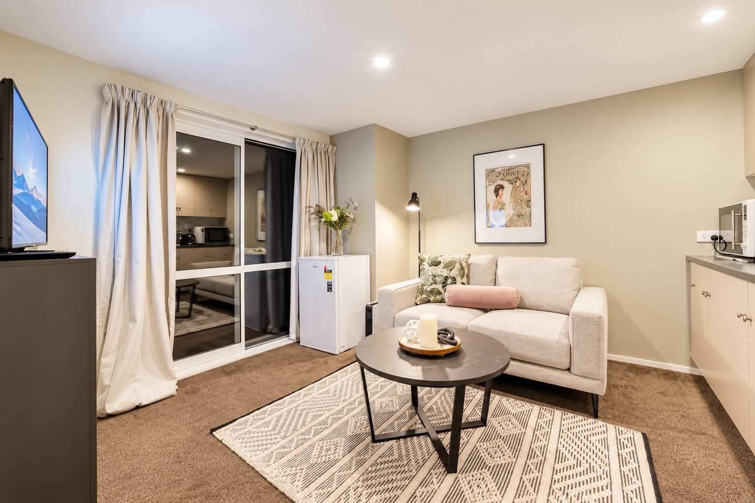 A modern living room with a sofa, a coffee table, a flat-screen TV, a refrigerator, a microwave, and a sliding glass door leading to a kitchen.