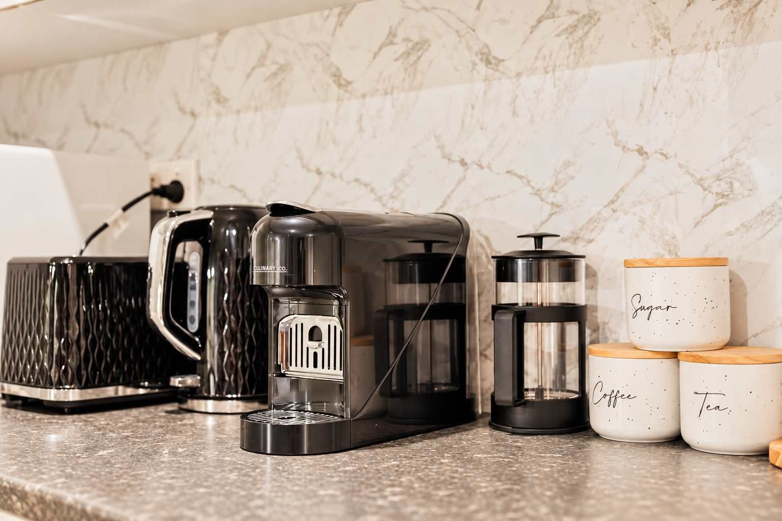 Eine Küchenarbeitsplatte mit einer Kaffeemaschine, einem Wasserkocher und drei Behältern, beschriftet mit 'Zucker', 'Kaffee' und 'Tee'.